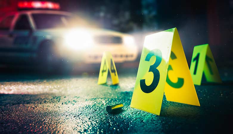 A bullet casing on the ground with evidence numbers around it and a police car in the background.