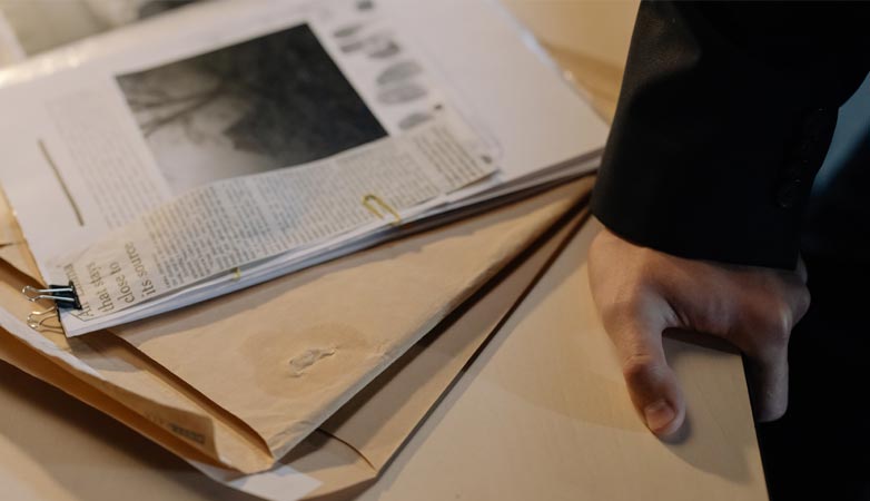 Close-up of a hand resting on a desk near a stack of investigative documents and newspaper clippings.