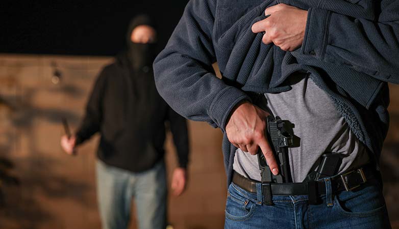 Someone pulling out a gun from their belt buckle as a man behind them approaches with a knife.